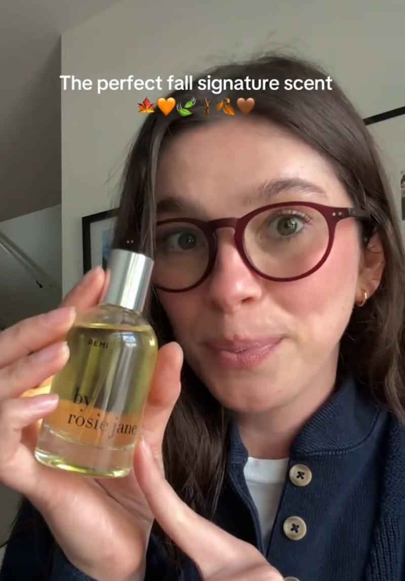 A UGC handheld demo where a woman holds up a perfume bottle towards the camera while smiling. Indoor casual setting with a neutral background.